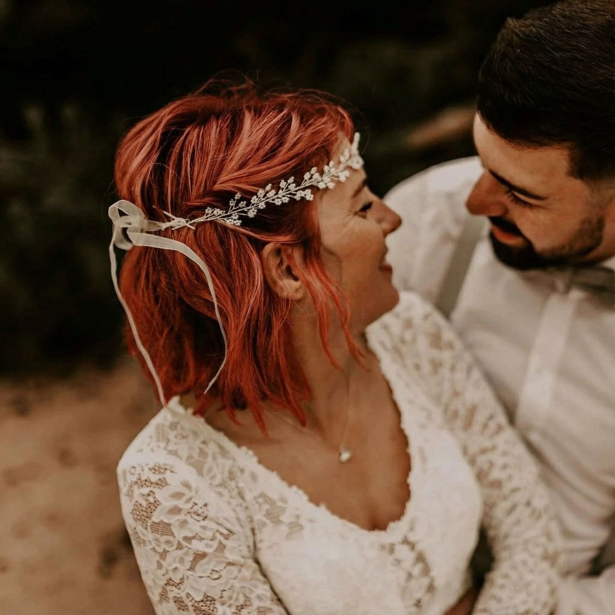 Eine Frau mit kurzen, gewellten roten Haaren, die mit einem weißen, geblümten Stirnband geschmückt sind, und ein Mann mit dunklen Haaren und einem Bart, beide in weißer Kleidung, sitzen im Freien dicht beieinander und lächeln sich liebevoll an. Die Frau trägt ein Spitzenkleid.