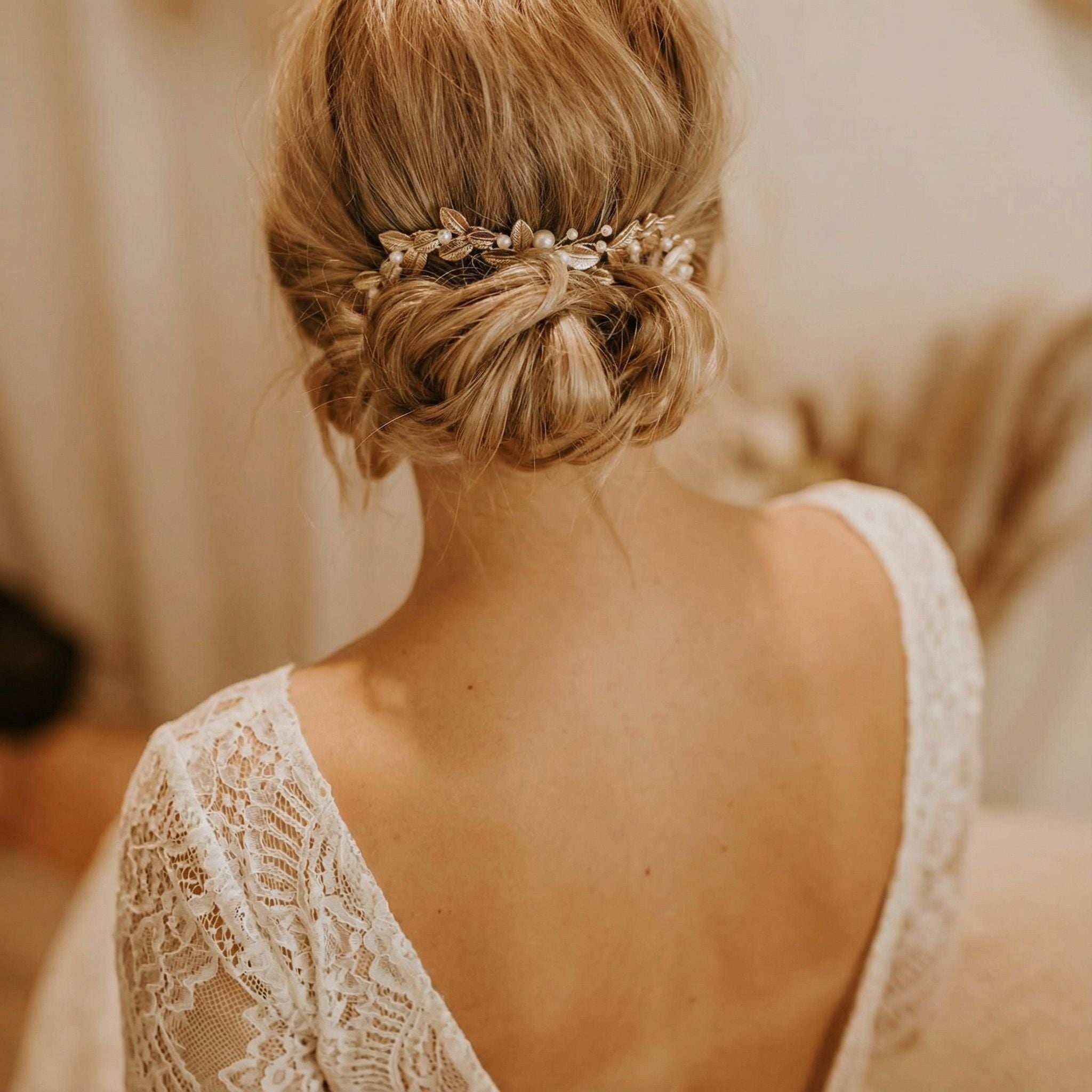 Eine Frau mit blondem Haar, das zu einem niedrigen, eleganten Dutt frisiert und mit einem zarten Haarschmuck aus Perlen und Blättern verziert ist. Sie trägt ein weißes Spitzenkleid mit offenem Rücken, und der Hintergrund ist mit neutralen Tönen sanft verschwommen.