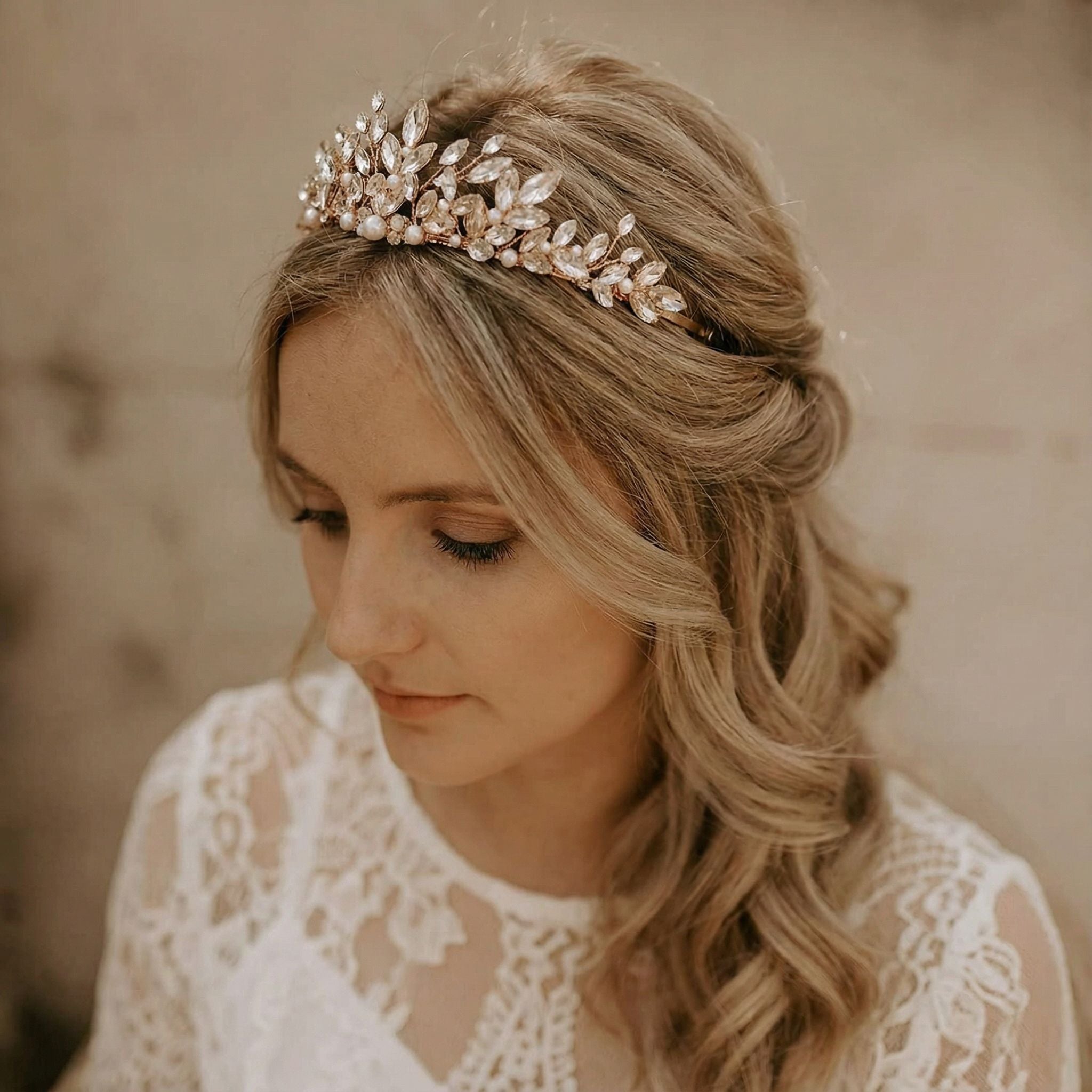 Eine Frau mit gewelltem blondem Haar, das halb hochgesteckt ist, trägt ein glitzerndes, verziertes Diadem mit Perlen und Kristallen. Sie ist in ein weißes Spitzenkleid gekleidet und blickt nachdenklich nach unten, mit einem weichen beigen Hintergrund im Hintergrund.