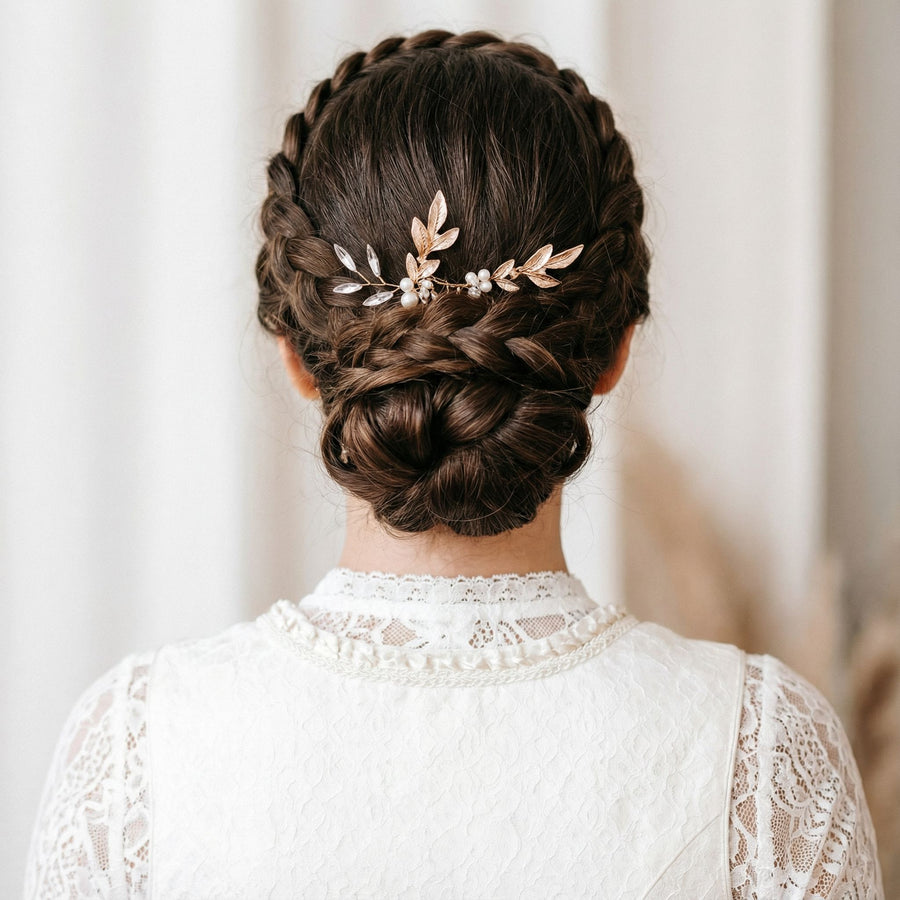 Eine Frau mit dunkelbraunem Haar, das zu einem geflochtenen Scheitel und einem niedrigen Dutt frisiert ist, geschmückt mit einem blattförmigen Haarschmuck aus Gold und Perlen. Sie trägt ein weißes Spitzenkleid, von hinten gesehen, mit neutralem, sanft verschwommenem Hintergrund.