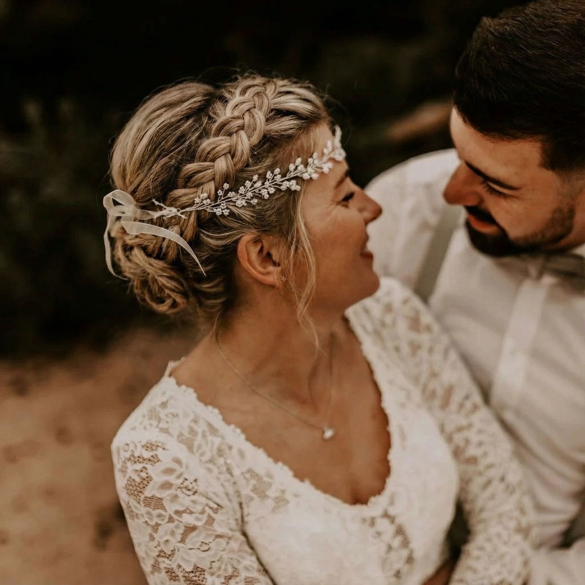 Eine Braut und ein Bräutigam lächeln sich gegenseitig an. Die Braut trägt ein Spitzenkleid, einen zarten Blumenkopfschmuck und ihr Haar zu einer geflochtenen Hochsteckfrisur. Der Bräutigam hat einen akkurat gestutzten Bart und trägt ein weißes Hemd mit Hosenträgern. Der Hintergrund besteht aus unscharfem Grün.