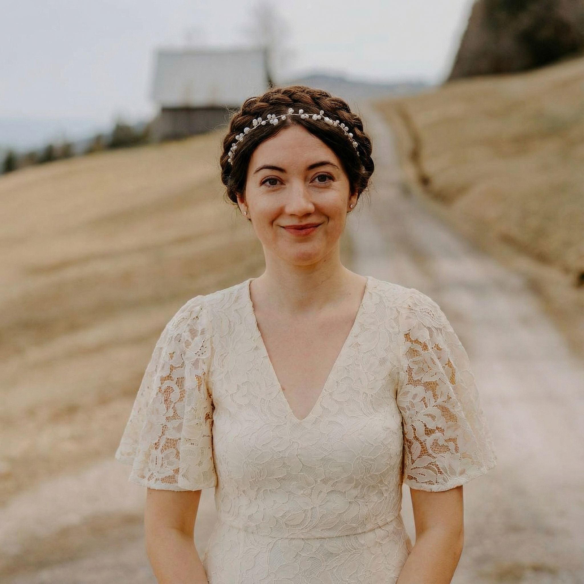Eine Frau in einem weißen Spitzenkleid steht auf einem Feldweg mit braunem Gras auf beiden Seiten. Sie hat braunes, geflochtenes Haar, das mit einem juwelenbesetzten Stirnband geschmückt ist, und lächelt sanft. Im Hintergrund sind ein hölzernes Gebäude und ferne Hügel unter einem wolkenverhangenen Himmel zu sehen.
