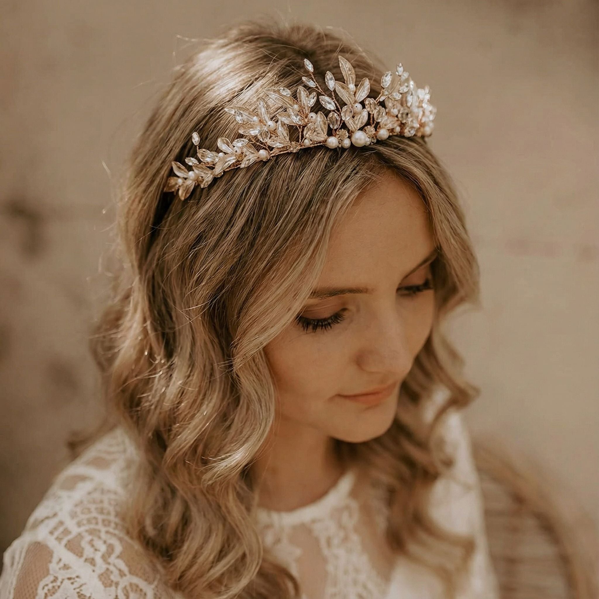 Eine Frau mit gewelltem, blondem Haar trägt ein goldenes, mit Perlen und Kristallen verziertes Diadem. Sie blickt sanft nach unten und ist in ein zartes weißes Spitzenkleid gekleidet, das einen warmen, neutralen Hintergrund hat. Die Stimmung ist sanft und elegant.