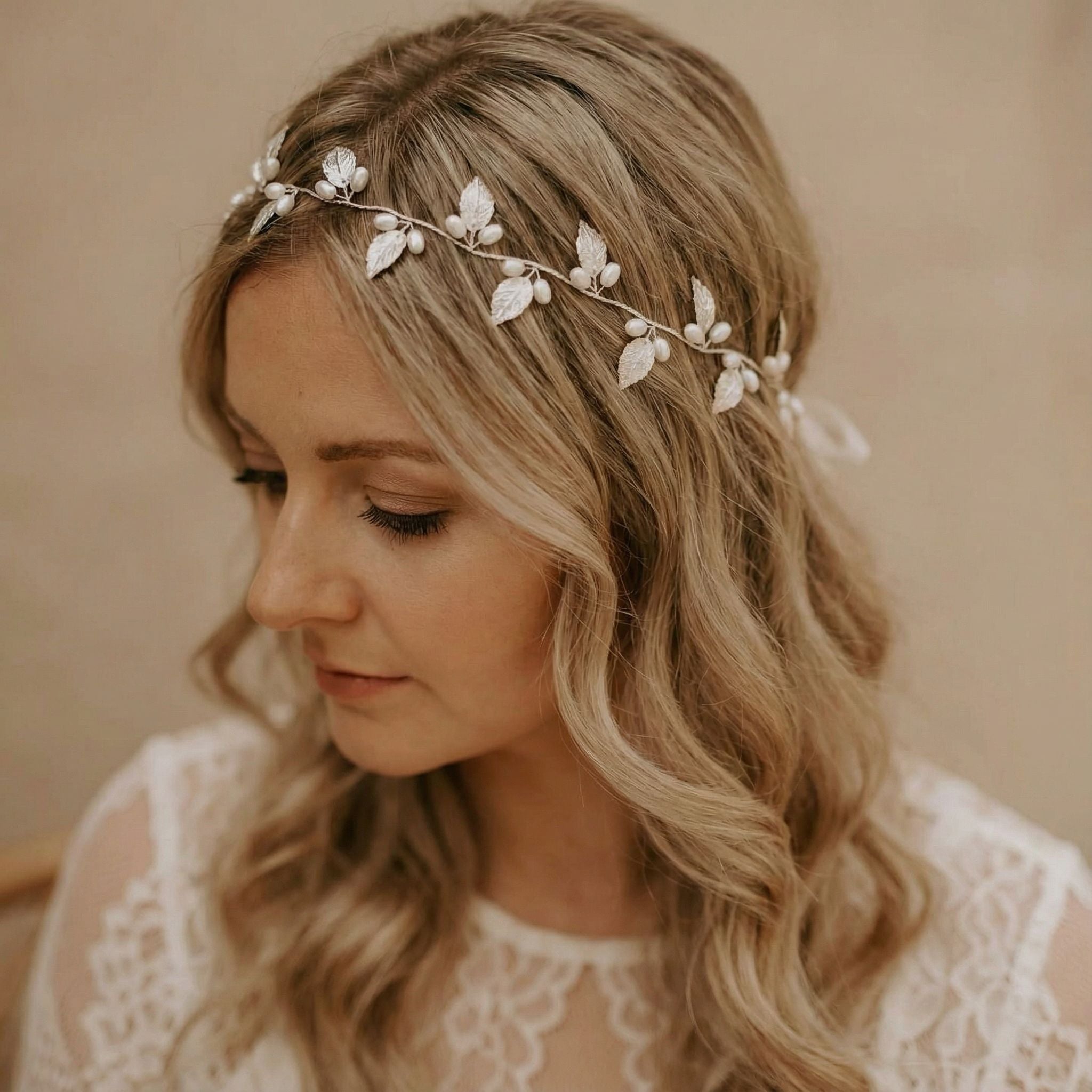 Eine Frau mit langem, gewelltem blondem Haar trägt eine zarte Kopfbedeckung, die mit weißen Blättern und Perlen verziert ist. Sie ist mit einem weißen Oberteil mit Spitzenmuster bekleidet und blickt nach unten, mit einem weichen, neutralen Hintergrund hinter ihr.