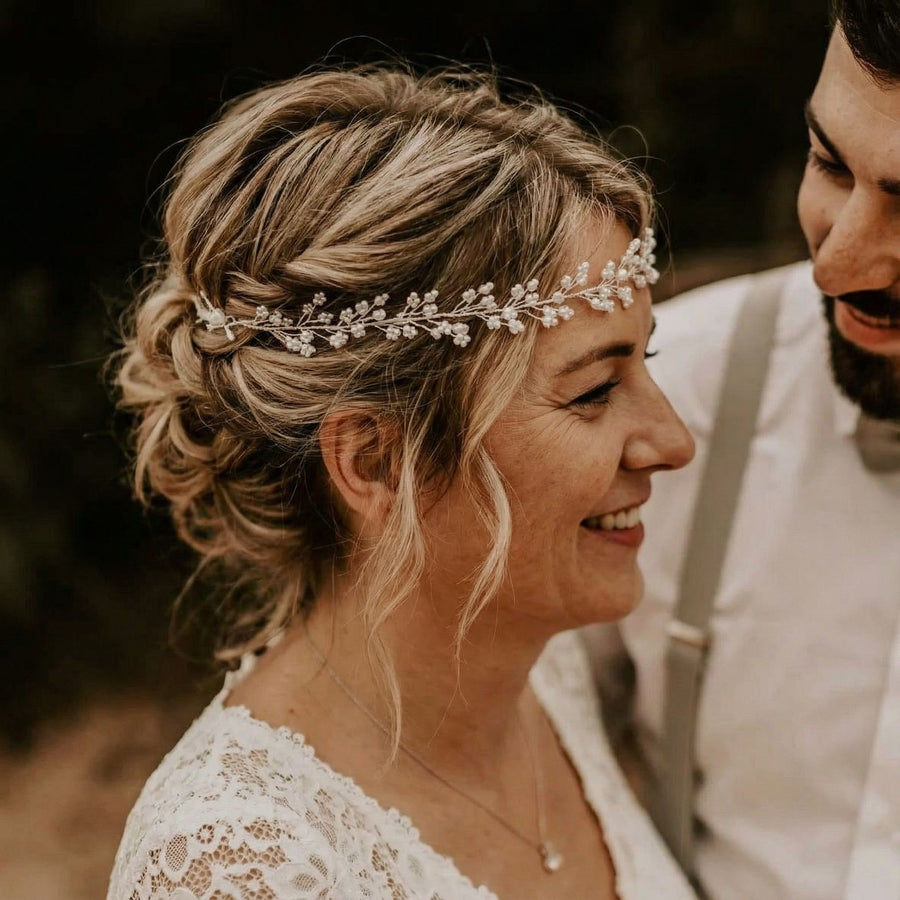 Eine Frau mit gewelltem Haar in einer eleganten Hochsteckfrisur, geschmückt mit einem zarten Perlenschmuck, lächelt einen bärtigen Mann neben sich an. Sie trägt ein weißes Spitzenkleid. Der Hintergrund ist unscharf und suggeriert eine natürliche Umgebung im Freien.