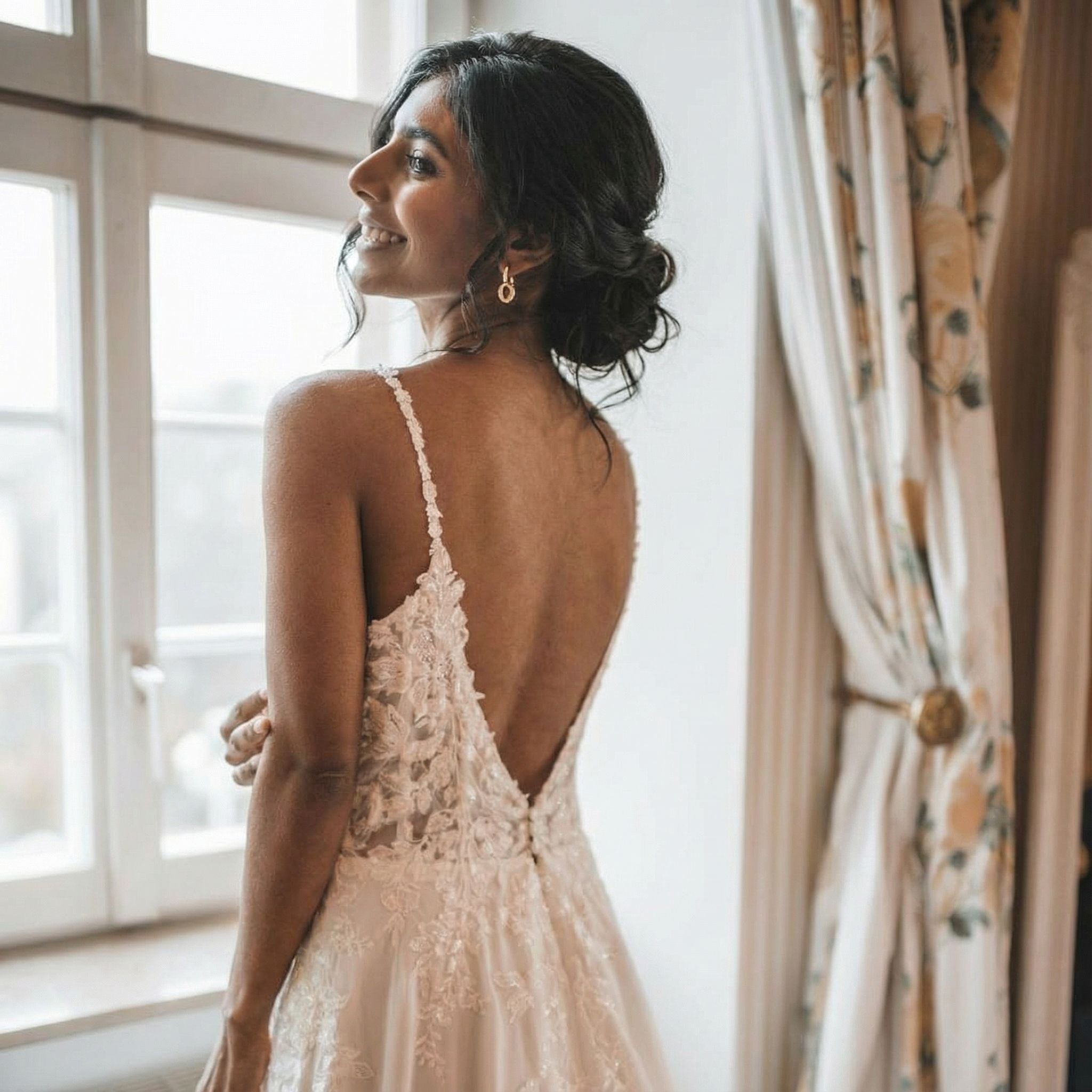 Eine Frau mit dunklem Haar in einer lockeren Hochsteckfrisur steht an einem Fenster und lächelt über ihre Schulter. Sie trägt ein ärmelloses, weißes Hochzeitskleid mit Spitzenrücken und Blumendetails. An ihren Ohren baumeln goldene Ohrringe. Cremefarbene Vorhänge mit einem Blumenmuster rahmen das Fenster ein.